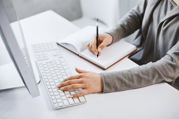 A focused young woman multitasking by typing on a keyboard and taking notes while studying business graphics on her computer screen, exemplifying productivity and dedication.